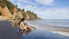 Donna seduta sulla spiaggia nera vicino al mare con scogliere sullo sfondo