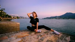 Donna rilassata seduta sul molo al tramonto con mare e montagna sullo sfondo